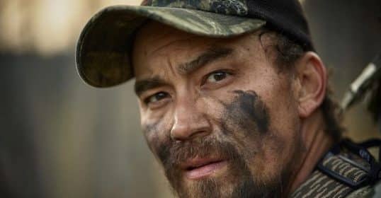 A close-up of a man with camouflage paint on his face, wearing military apparel, and looking off-camera during Podcast #902 on