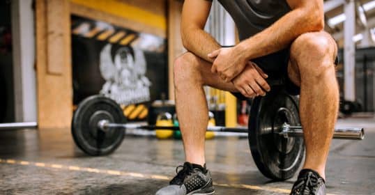 A person resting with hands on knees in a gym, with a barbell on the floor nearby, as if they don't feel like working out.