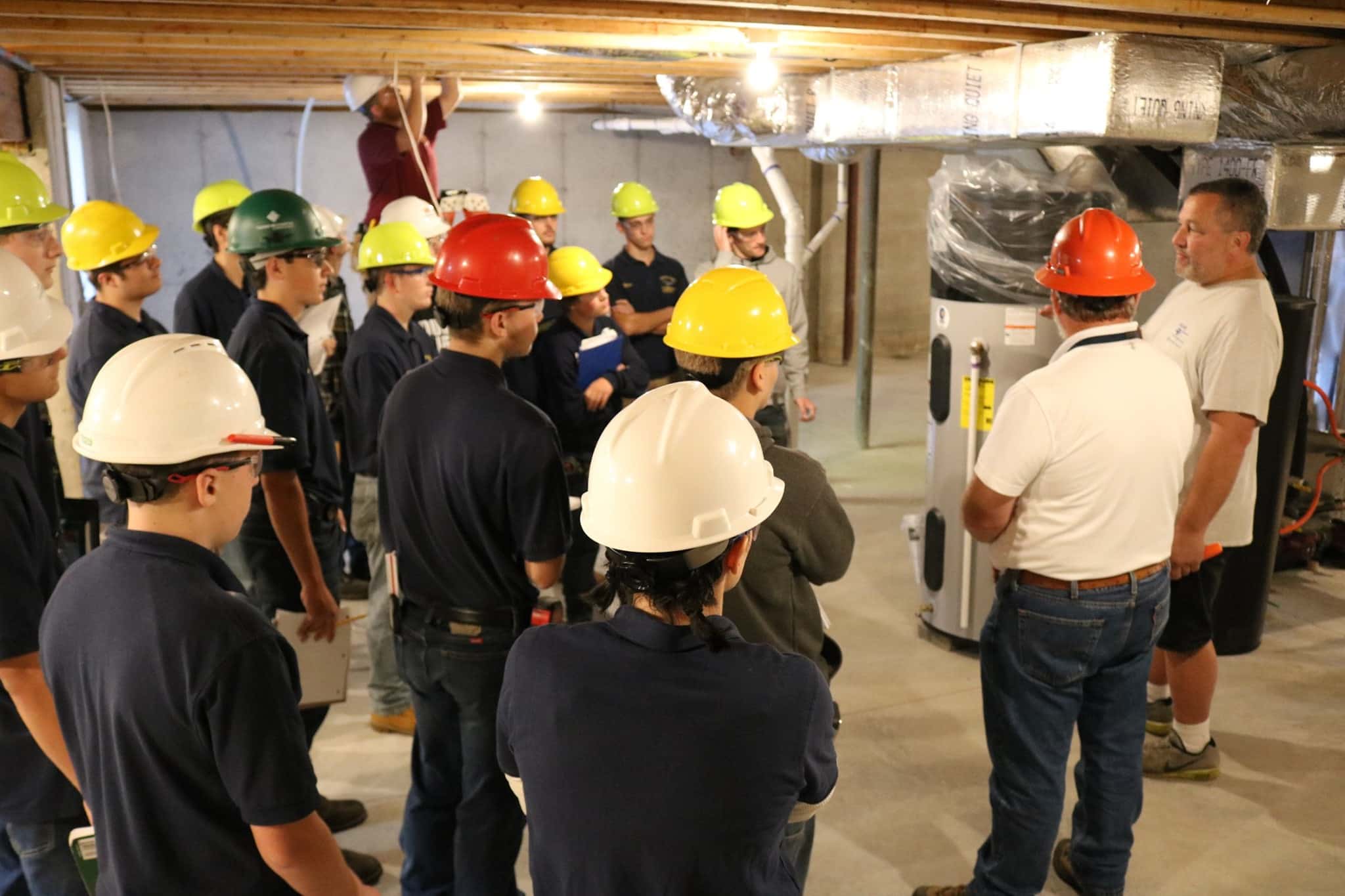 Group of construction trainees in hard hats receiving Trades School instruction in a building's basement.