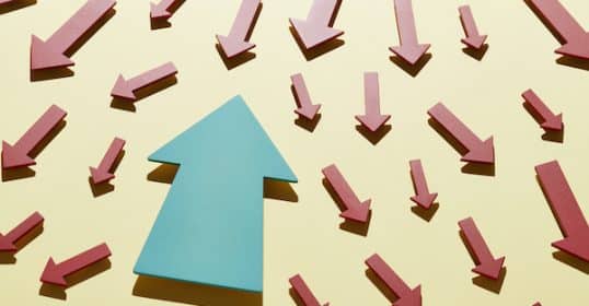 A blue arrow pointing upwards amidst numerous red arrows pointing downwards against a yellow background, symbolizing anti-mimetic life choices.