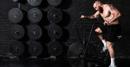 A man with tattoos working out on an assault bike in a gym with weights in the background.