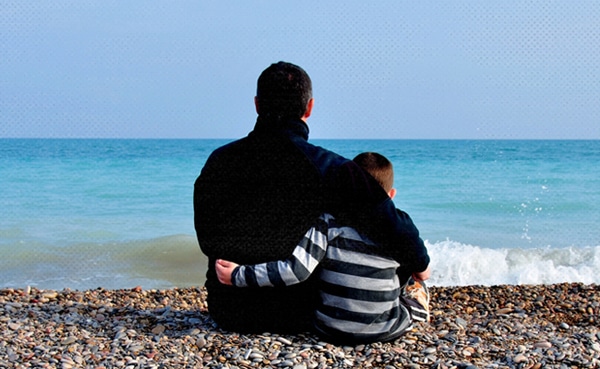 An adult and a child, demonstrating the essence of parenting, sitting close together on a pebble beach, looking out at the sea.