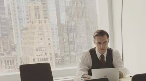Business professional working on a laptop in an office with a city view, listening to podcast advice on making love last.