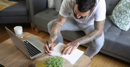 A man with tattoos writing in a notebook at a coffee table with a laptop and a cup of coffee, in a cozy living room, enjoying his Sunday habits.