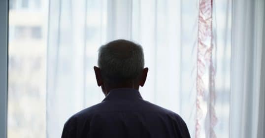 A person, getting old, seen from behind looking out of a window with sheer curtains.