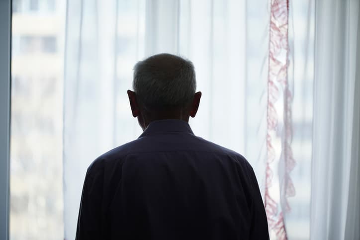 A person, getting old, seen from behind looking out of a window with sheer curtains.