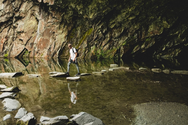 Person stepping on rocks across a shallow water body inside a rocky gorge, contemplating the "5 Shifts of Manhood" discussed in Podcast #926.
