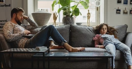 A man and a young girl lounging on a couch in a living room, both appearing relaxed and pensive, possibly discussing the cure for boredom.