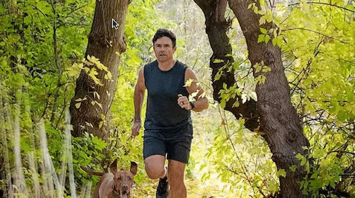 Man jogging on a forest path, listening to a podcast, with his dog.