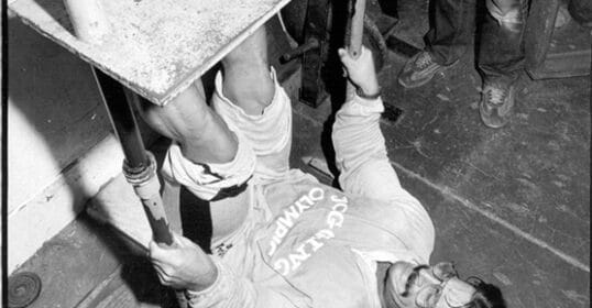 A man in athletic clothing lies on his back using a homemade gym apparatus, gripping bars and lifting a weighted platform with his legs in a pose reminiscent of a classic Mike Mentzer workout.