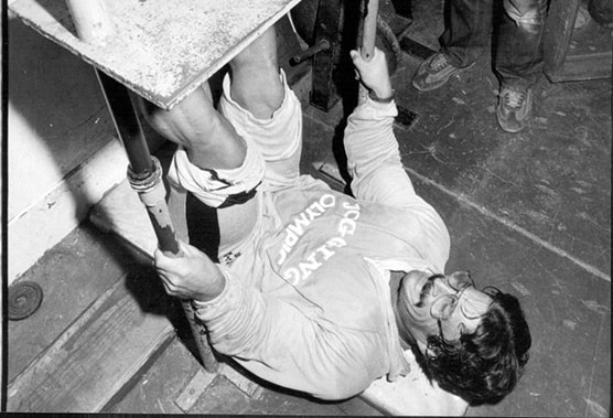 A man in athletic clothing lies on his back using a homemade gym apparatus, gripping bars and lifting a weighted platform with his legs in a pose reminiscent of a classic Mike Mentzer workout.