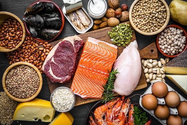 Various protein-rich foods such as meats, fish, legumes, eggs, and nuts arranged on a dark surface support the Protein Leverage Hypothesis.