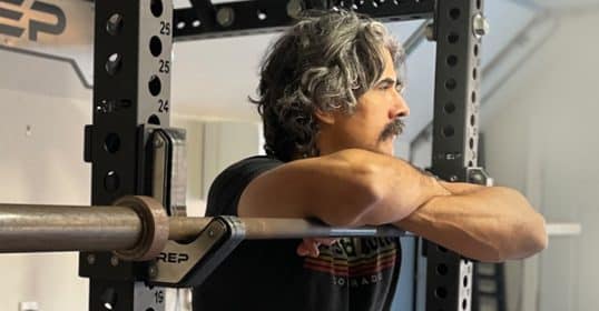 Man with gray hair resting arms on a fitness barbell in a squat rack.