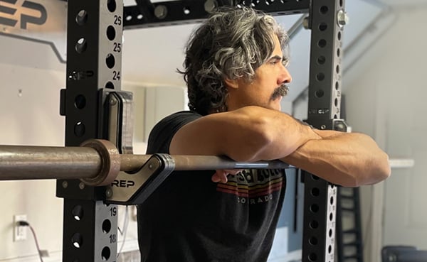 Man with gray hair resting arms on a fitness barbell in a squat rack.