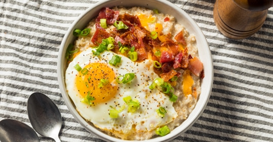 A bowl of savory oatmeal topped with a fried egg, bacon, and chopped green onions.