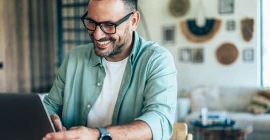 Man smiling while working on a laptop at a home office, boosting his productivity.