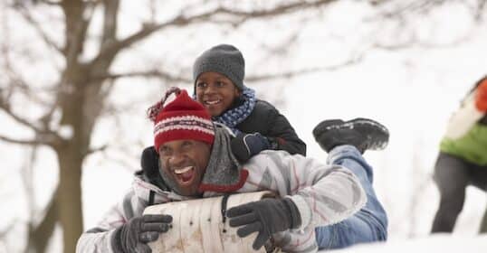 A joyful child rides piggyback on an adult while sledding down a snowy hill, embodying the theme of