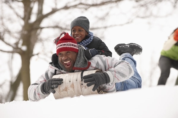 A joyful child rides piggyback on an adult while sledding down a snowy hill, embodying the theme of "Having More Fun".