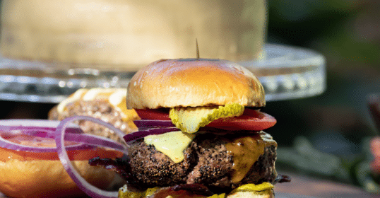 A gourmet cheeseburger with pickles, tomatoes, red onions, and lettuce served on a wooden board as part of Grandaddy's Sunday Dinner.