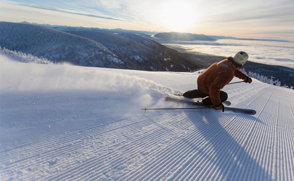A skier in an orange jacket carving a careful turn on a groomed slope with a scenic mountain backdrop at sunset.