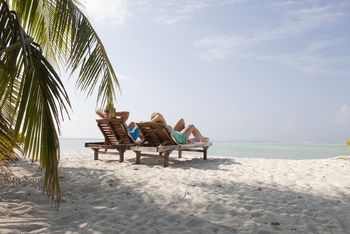 Two people relaxing on sun loungers by the beach under clear skies during their vacation.