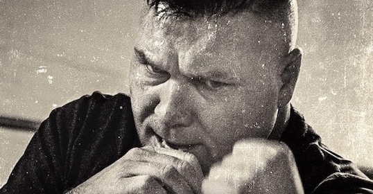 Black and white image of an intense man clenching his fist, facing downward in a gritty, textured close-up from Podcast #985.