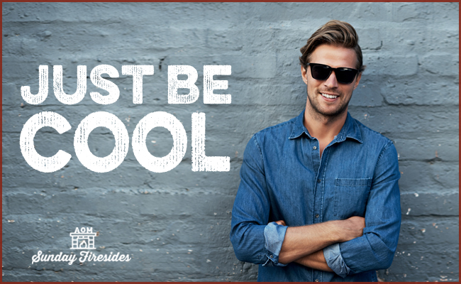 Smiling man in sunglasses leaning against a blue wall with the phrase "Just Be Cool" painted above his head.