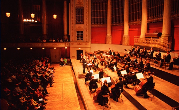 Orchestra performing classical music on stage in a grand hall with an audience in the foreground.