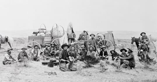 Black and white historical photo of a group of people in western attire, including cowboys, sitting and standing around covered wagons on a grassy plain.