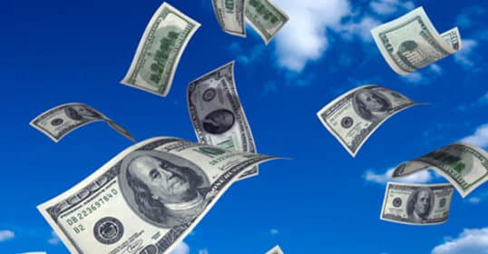 U.S. hundred dollar bills fluttering against a clear blue sky with white clouds symbolize a sudden financial windfall.