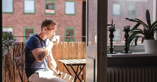A man sitting at a small outdoor table embraces his everyday rituals by drinking from a cup on a sunny balcony, with green plants and a decorative item visible inside.