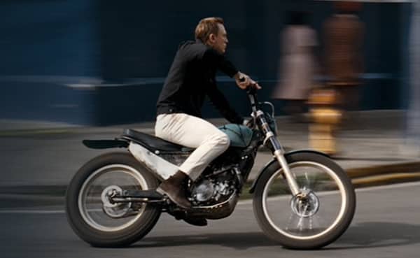 A man in white jeans and casual attire riding a motorcycle swiftly on an urban street, with motion blur indicating speed.