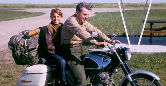 Two people on a motorcycle: a child smiling at the back and an older person driving. The motorcycle, well-maintained and reliable, has bags strapped to the back. They are in an open area with grass and a picnic table in the background, embodying a moment of simple zen-like joy.
