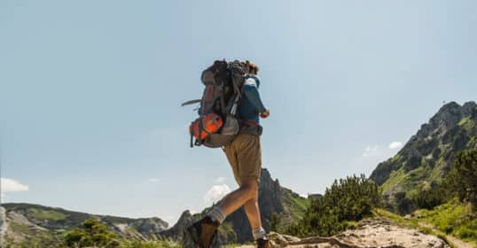 A person with a backpack hikes up a trail in a mountainous landscape under a clear sky, enjoying their first backpacking trip.