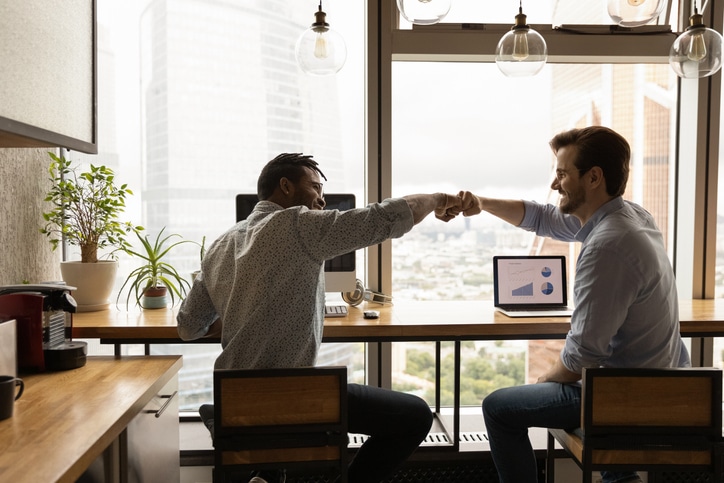 Two individuals sit at a desk in an office, fist-bumping. A laptop displaying a pie chart and bar graph is open in the background, alongside plants and large windows overlooking city buildings.