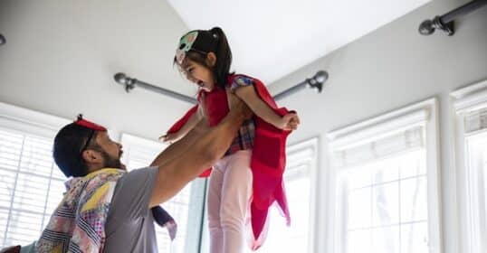 A dad is lifting a young child in the air. Both are wearing capes and masks, playing in a room with large windows. He embraces his essential role, making kids awesome through imaginative play.