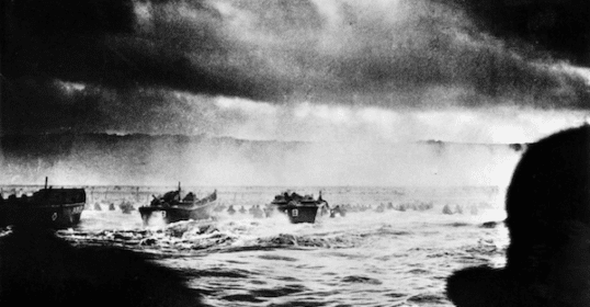 Black and white photo of WWII soldiers wading through water towards the beach during the D-Day invasion, with landing crafts in the background and dark, cloudy skies overhead. 80 Years Later we revisit this moment in Podcast #996.