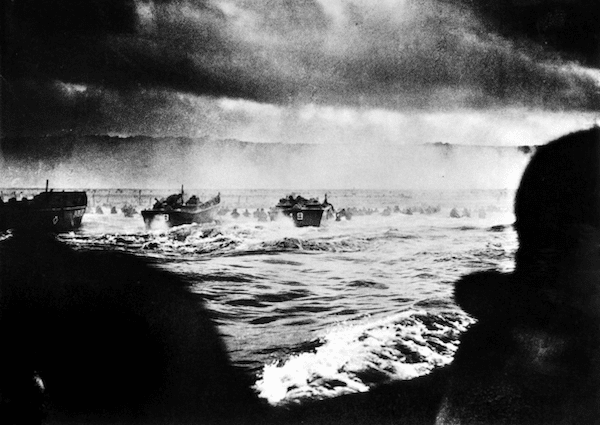 Black and white photo of WWII soldiers wading through water towards the beach during the D-Day invasion, with landing crafts in the background and dark, cloudy skies overhead. 80 Years Later we revisit this moment in Podcast #996.
