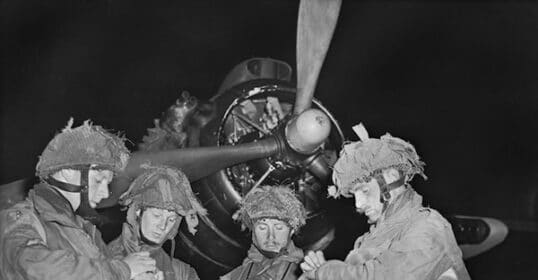 Four soldiers wearing helmets and military gear check their watches in front of a large aircraft propeller at night. This scene, reminiscent of D-Day, captures one of those rarely-seen photographs that offer a haunting glimpse into wartime preparations.