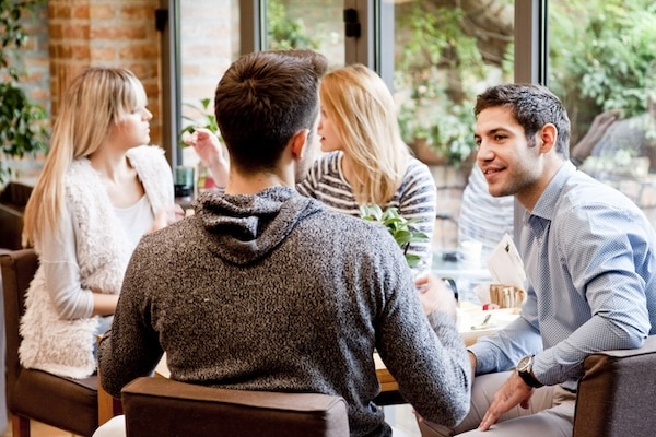 Four people sit at a table in a café, engaged in conversation. Two men, showcasing the nuances of male friendships, face the camera, while two women are seen in profile. The background reveals a bright, greenery-filled outdoor area.