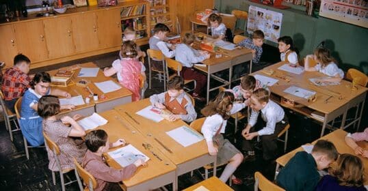 Children are seated at tables in a classroom, engaging in various activities and conversations with each other. Some are drawing on papers while others discuss their thoughts or look around. Bookshelves filled with resources on moral education stand proudly in the background.