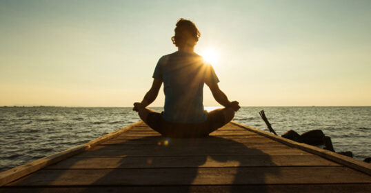 A person sits cross-legged and meditates on a wooden pier overlooking a body of water at sunset during Sunday Firesides.
