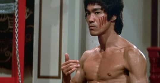 A shirtless man with claw marks on his chest and face stands indoors, gesturing with his hand and looking determined, embodying the intensity of a Bruce Lee workout.