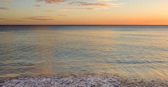 Sunset over a calm ocean wave gently lapping onto a sandy shore, reminiscent of a peaceful Sunday Firesides. The sky displays a gradient of colors from orange to blue.