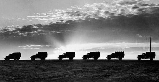 Silhouetted trucks drive in line along a ridge at sunset, demonstrating precise convoy execution against a backdrop of a cloudy sky and beams of sunlight.