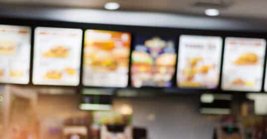 Blurry image of a fast food restaurant counter with illuminated menu boards offering meals less than 600 calories and high in protein, and a person using a phone in the foreground.