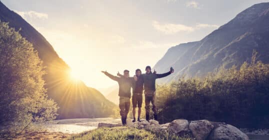 Three people stand together with arms raised in celebration on a rocky riverbank, surrounded by lush mountains, with the sun setting behind them—an ideal scene of enduring friendships.