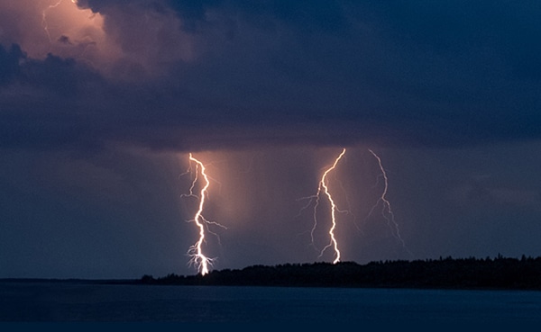 Two bolts of lightning strike a dark, cloudy sky over a body of water near a forested shoreline during nighttime, reminiscent of past summers spent around Sunday Firesides.