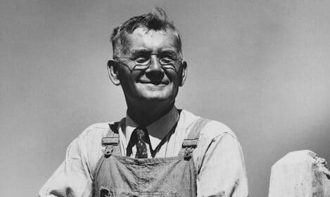 Black and white photo of an older man wearing glasses, a tie, and overalls. He is smiling with arms crossed in front of him against a clear 1930s sky.