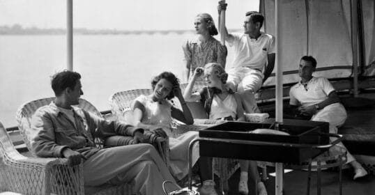 A group of people dressed in casual clothing relax and converse on an outdoor deck with wicker furniture, magazines, and a small cart. The background shows a body of water, creating a personal atmosphere perfect for unwinding.